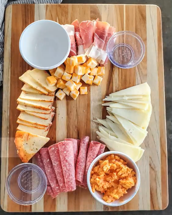 A charcuterie board with two plastic bowls and two ceramic bowls, and cheese slices laid onto the left side and right side of the board, and sliced meat onto the top and bottom side of the board. 
