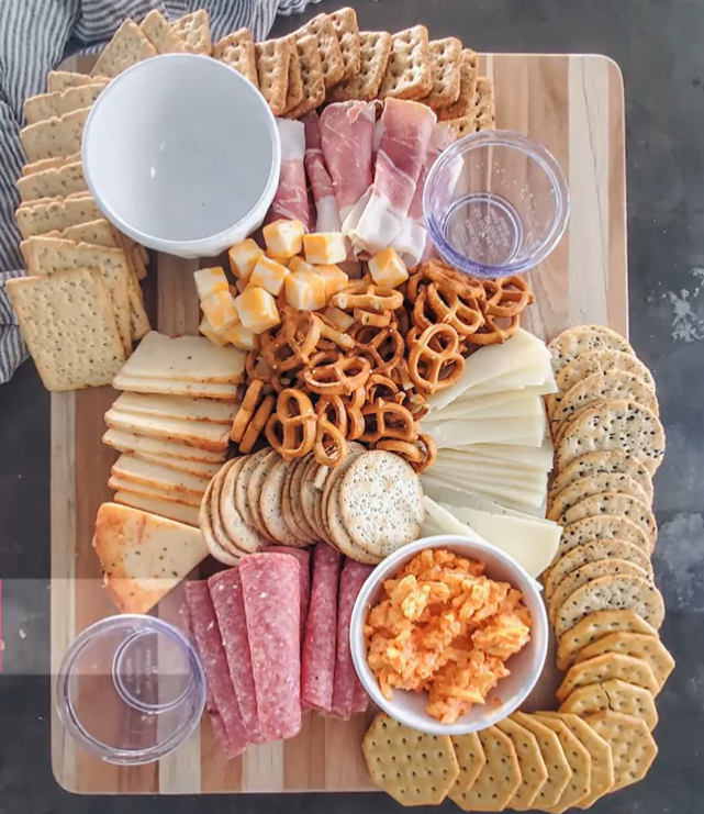 A charcuterie board with cheese slices, sliced meat, and four bowls, as well as crackers on the of the board.
