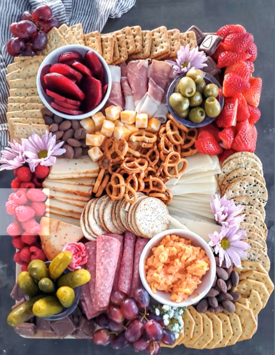A charcuterie board with cheese slices, sliced meat, and four bowls, as well as crackers on the of the board, and fruits on the edges of the board