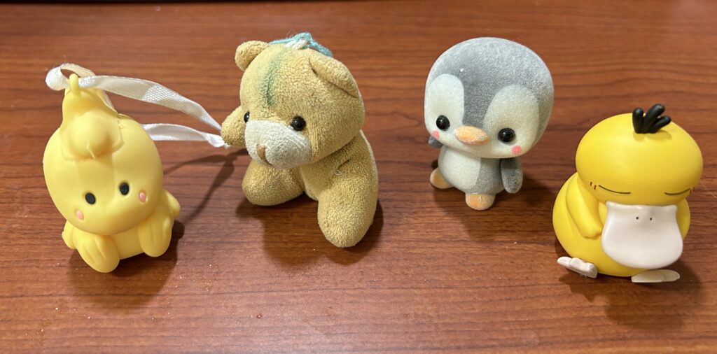 Four small toys, imaginary companions, on a wooden desk: a yellow chick with a ribbon, a plush bear, a penguin, and a Pokémon called Pysduck.