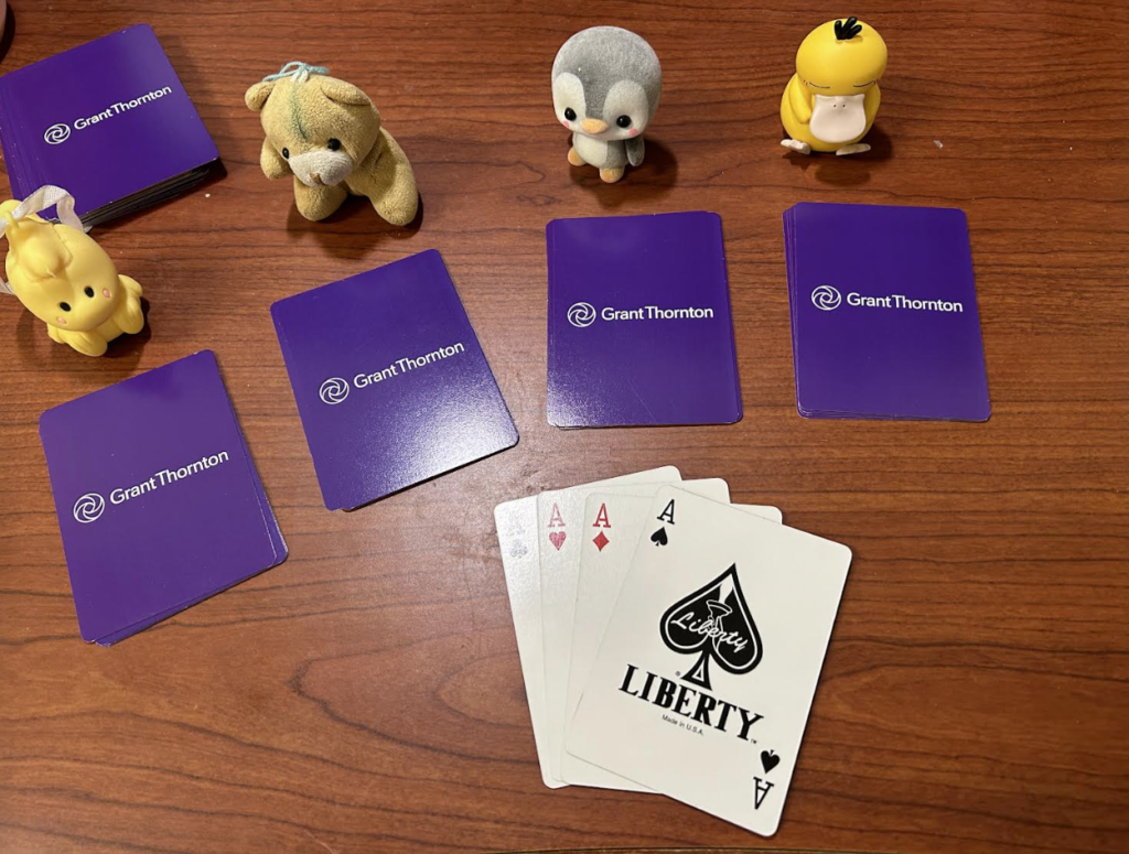 The cards are now distributed by a sample amount. The rest of the deck is in the background, and there are now the small toys placed by four of the distributed piles. This time the fifth deck is flipped over, showing all four of the cards as the four aces in the deck. 