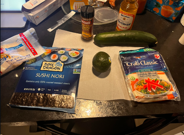 Ingredients for sushi roll: Japanese rice, toasted sesame seeds, avocado, cucumber, cutting board, parchment paper or sushi rolling mat, rice cooker, and imitation crab sticks