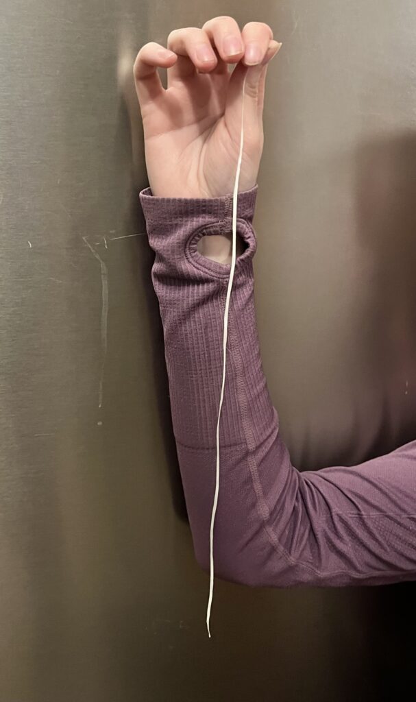 A hand is holding up a piece of floss approximately the length of the person's forearm.  The person is wearing a purple long sleeve shirt and the arm is in front of a gray background.