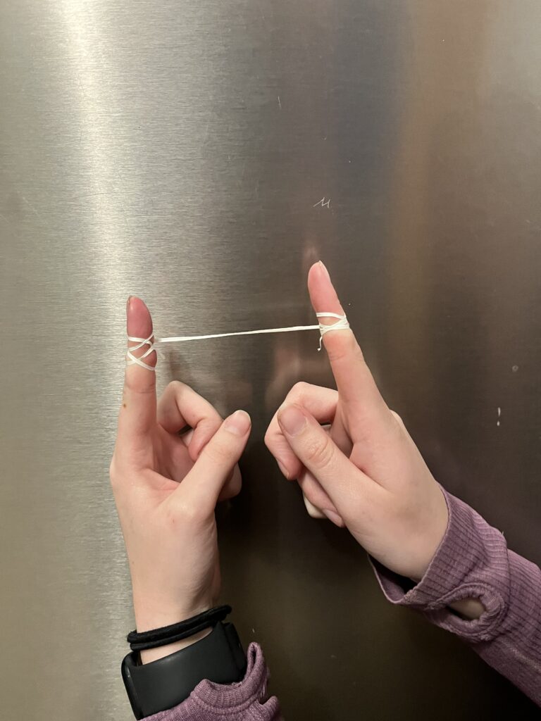 A person is holding up their hands with each index finger extended. Around each index finger, a piece of white floss is wrapped around and extended between the two fingers