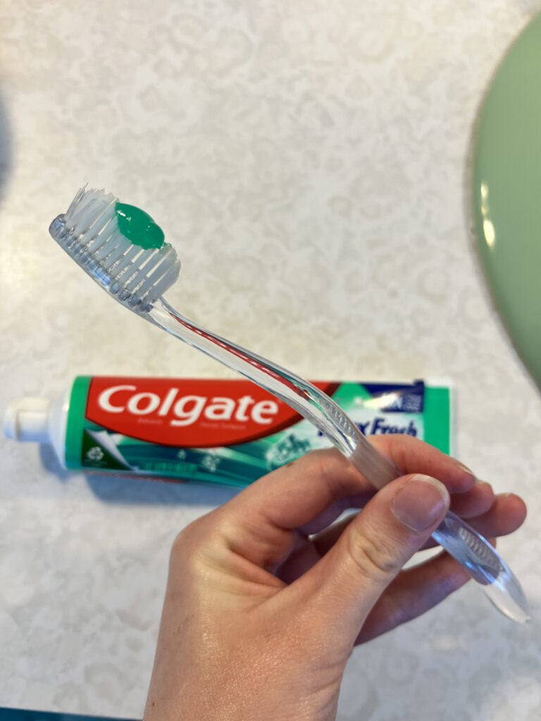 In the foreground, a toothbrush is being held with a pea sized amount of green toothpaste on it.  In the background, there is a white countertop with a tube of Colgate toothpaste resting on it.